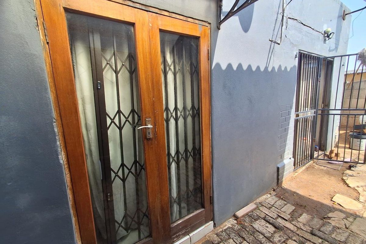 Rear exterior view showing wooden double doors with security gates, brick paving, and an awning attached to a painted masonry wall.