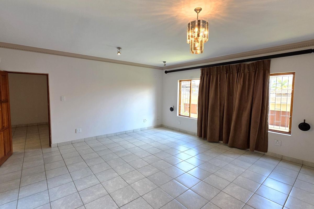 A spacious, tiled living room featuring neutral walls, decorative crown molding, a central chandelier, and large windows with curtain rods.