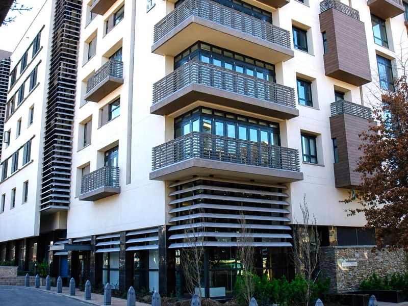 Modern multi-story apartment building facade featuring stacked balconies, metal louvred sun screens, and stone cladding accents at the ground level entrance.