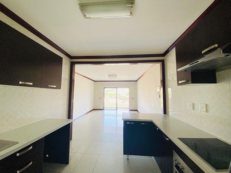 Open-plan kitchen featuring dark cabinetry, light countertops, and integrated appliances, flowing into a bright living area with access to a balcony.