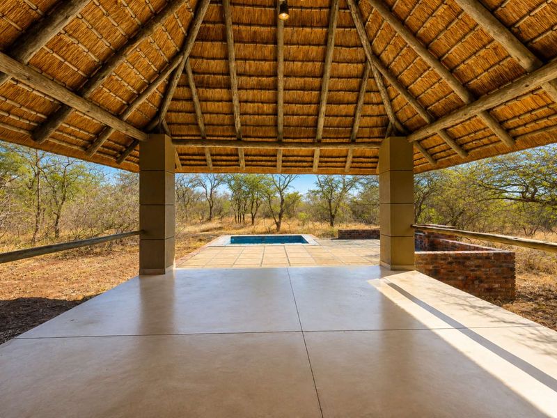 A spacious covered patio featuring a traditional thatched roof, tiled flooring, and direct access to an outdoor swimming pool set in a natural bushveld environment.