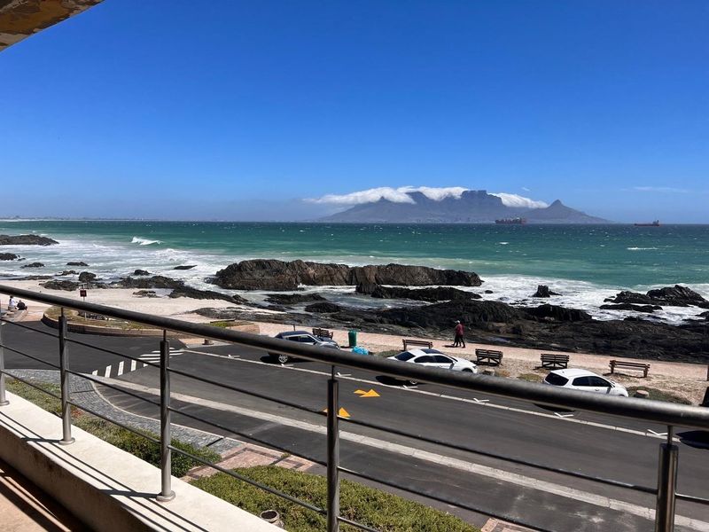 A scenic coastal view overlooking the ocean, rocky shoreline, and Table Mountain, framed by a modern stainless steel balcony railing.