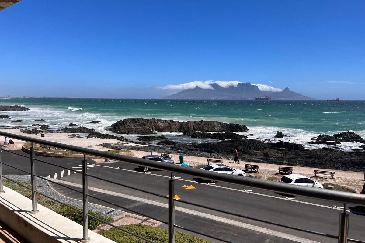 A scenic coastal view overlooking the ocean, rocky shoreline, and Table Mountain, framed by a modern stainless steel balcony railing.