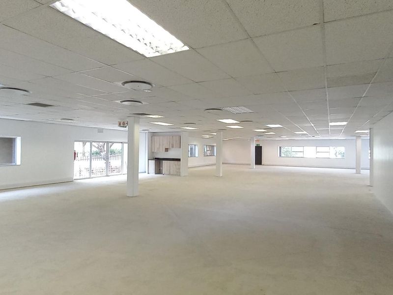 A spacious, open-plan office area featuring a suspended ceiling, recessed lighting, support columns, and a small built-in kitchenette, presented in a clean, vacant state.