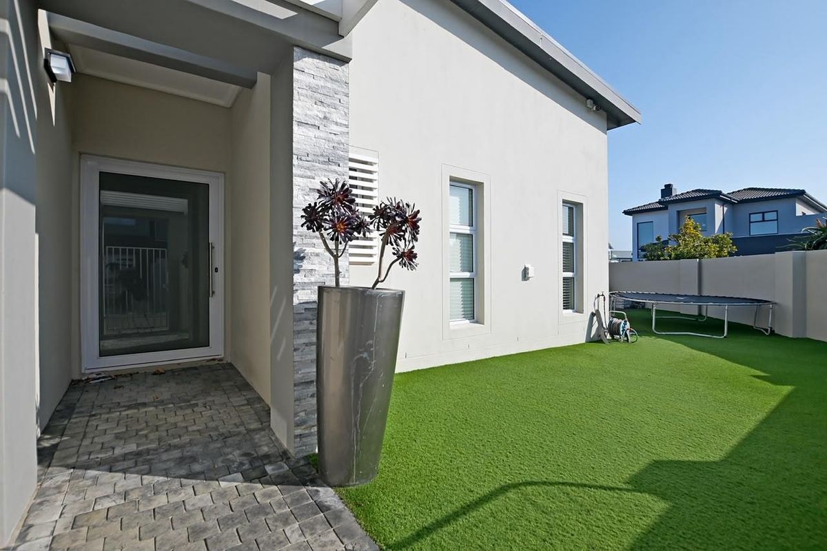 A well-maintained rear exterior featuring a paved walkway, artificial lawn, and stone-clad pillar leading to a secure entrance.