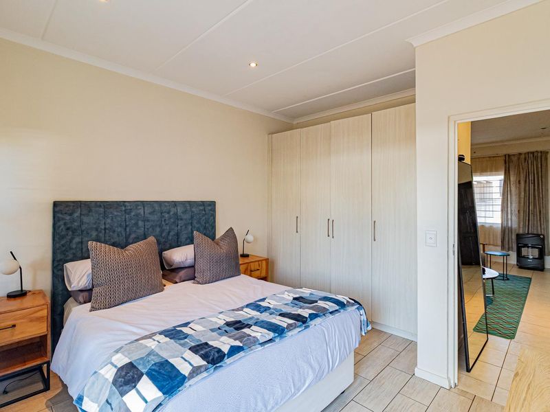 A well-maintained bedroom featuring built-in cupboards, tiled flooring, and recessed downlighting, with an adjoining view into the living area.
