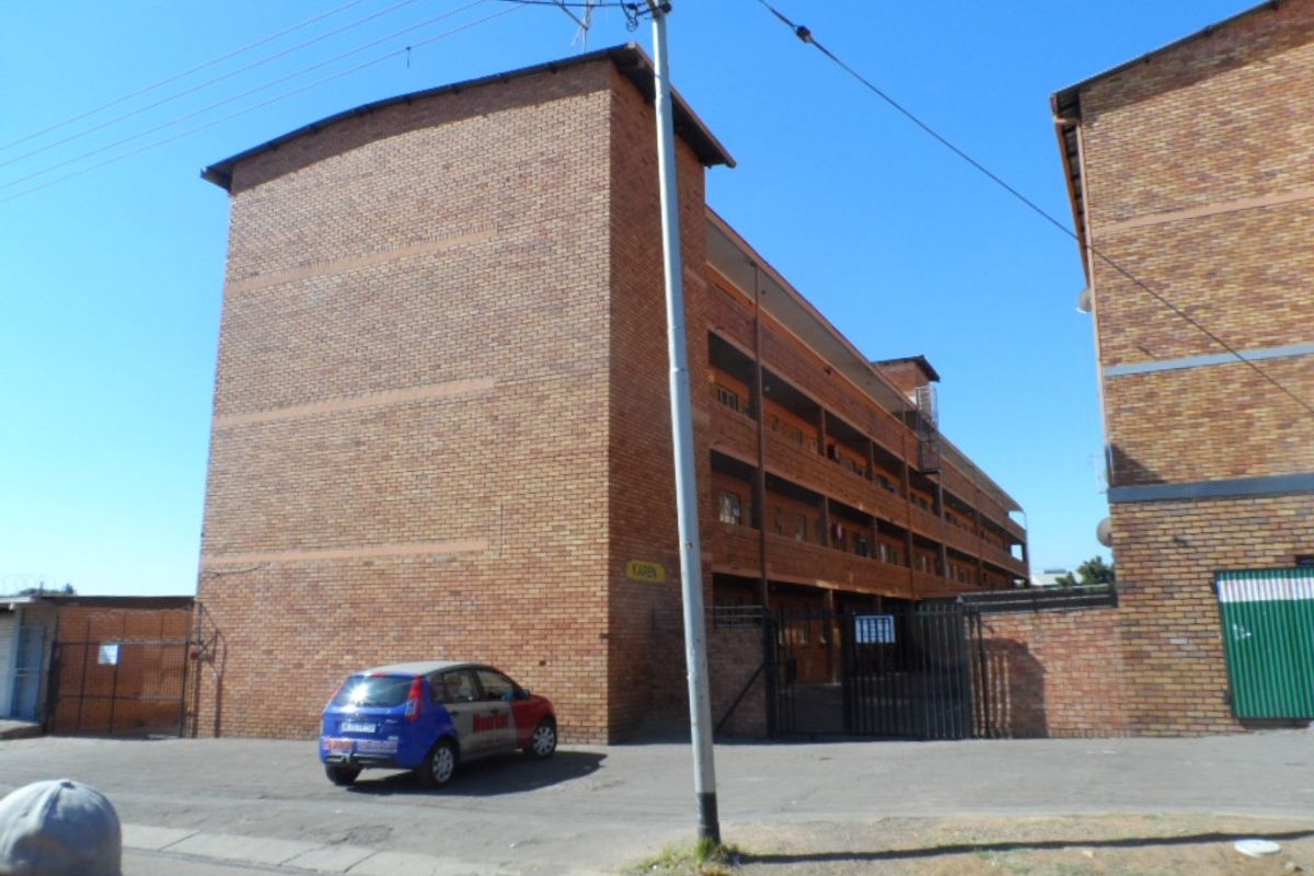 Exterior view of a multi-story face brick apartment building with a gated entrance and paved driveway area.