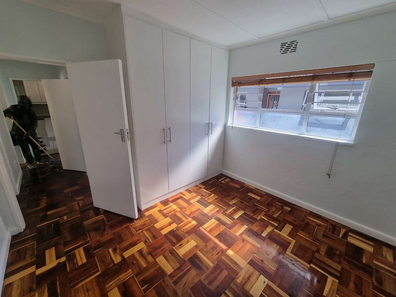 A bright, empty bedroom featuring polished parquet flooring, white built-in cupboards, and a window with wooden venetian blinds.