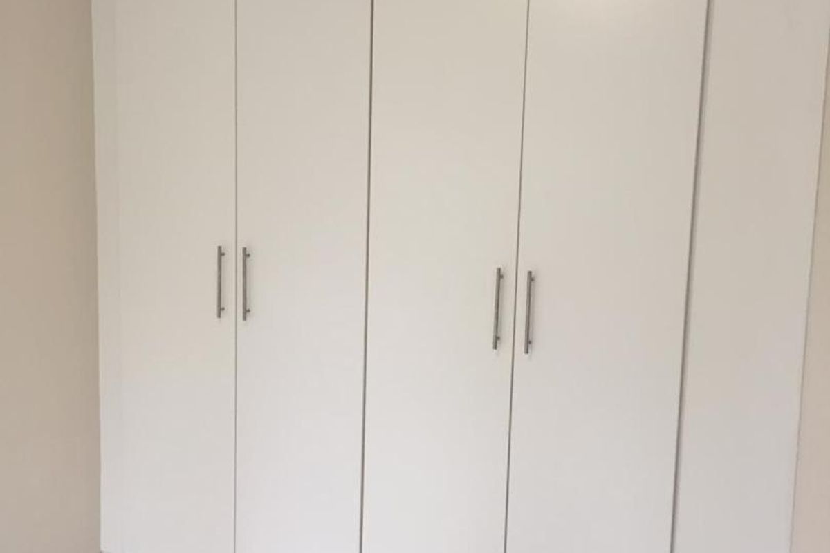 A clean, empty bedroom featuring neutral tiled flooring and a large, modern white built-in wardrobe with sleek metal handles.