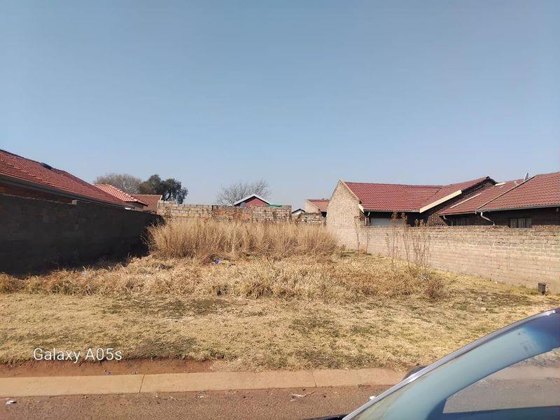 A vacant residential plot enclosed by perimeter masonry walls, featuring overgrown grass and weeds, situated in a developed neighborhood.