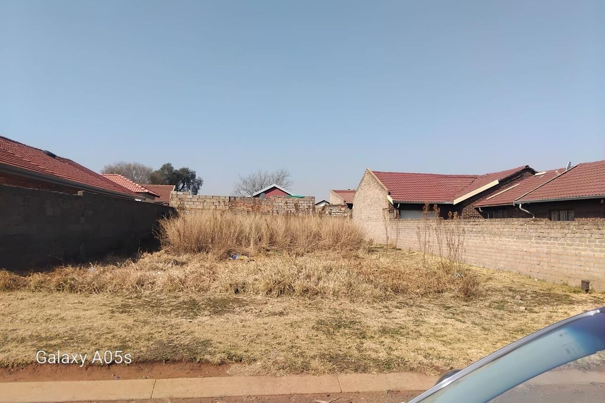 A vacant residential plot enclosed by perimeter masonry walls, featuring overgrown grass and weeds, situated in a developed neighborhood.