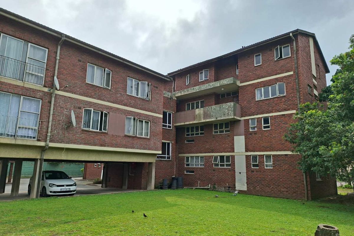 Rear view of a multi-story brick apartment building featuring concrete balconies, aluminium windows, and a shared ground-level lawn area.