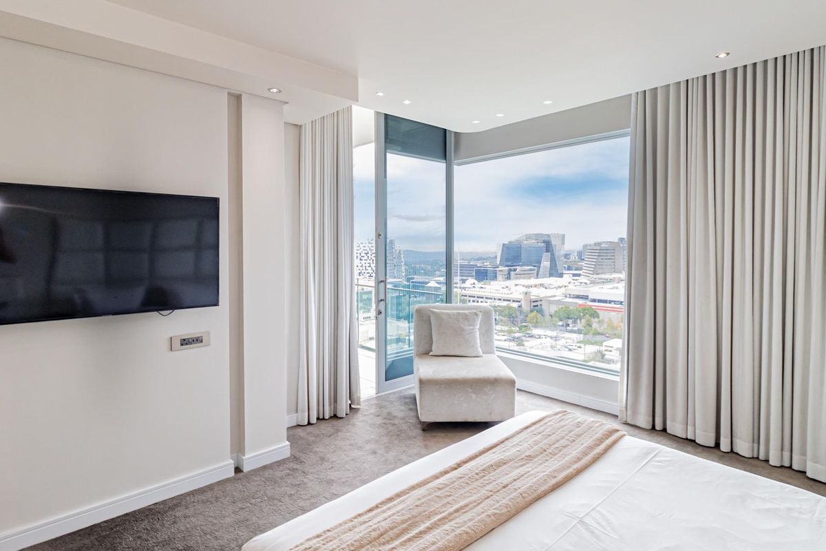 Modern bedroom featuring floor-to-ceiling windows with city views, integrated curtain tracks, recessed lighting, and direct access to a private balcony.