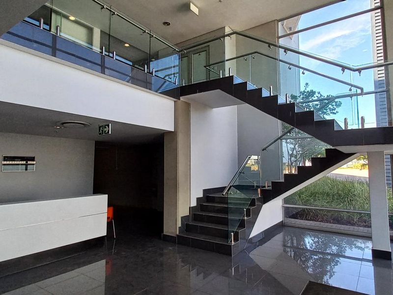 Modern industrial entrance lobby featuring a double-volume ceiling, glass balustrade staircase, polished floor tiles, and expansive floor-to-ceiling windows providing natural light.