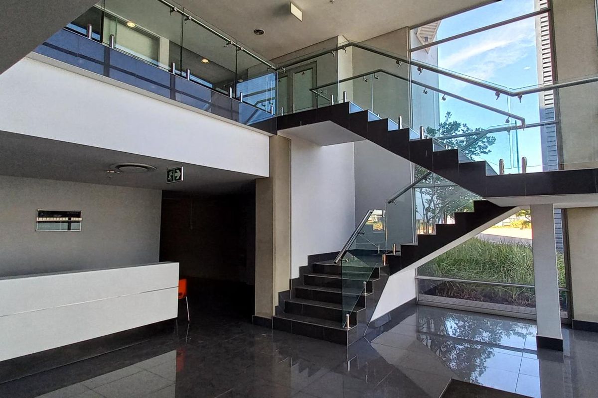 Modern industrial entrance lobby featuring a double-volume ceiling, glass balustrade staircase, polished floor tiles, and expansive floor-to-ceiling windows providing natural light.