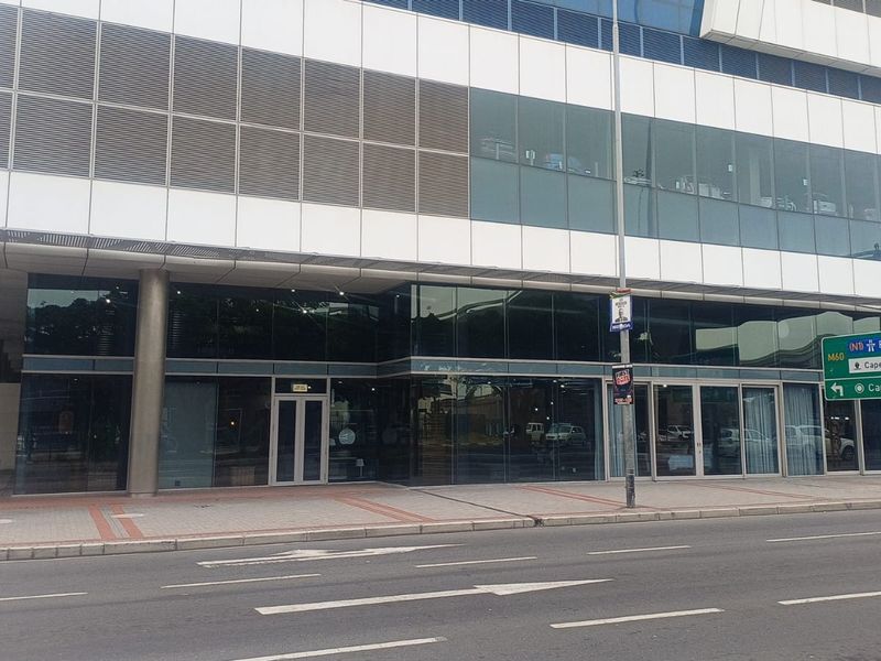 Modern commercial building facade featuring extensive glass curtain walling, metal cladding, and ground-level glass entrance doors situated along a paved street.