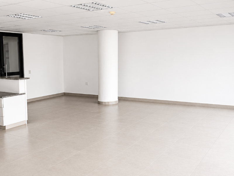 A bright, open-plan office space featuring neutral ceramic floor tiles, a suspended ceiling with recessed lighting, and a central structural column.