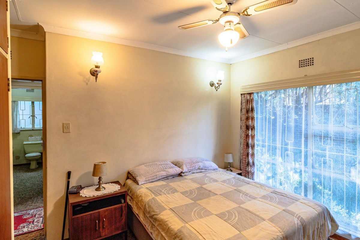 A bedroom featuring a ceiling fan, wall-mounted light fixtures, and a large window with security bars, with an adjoining en-suite bathroom.
