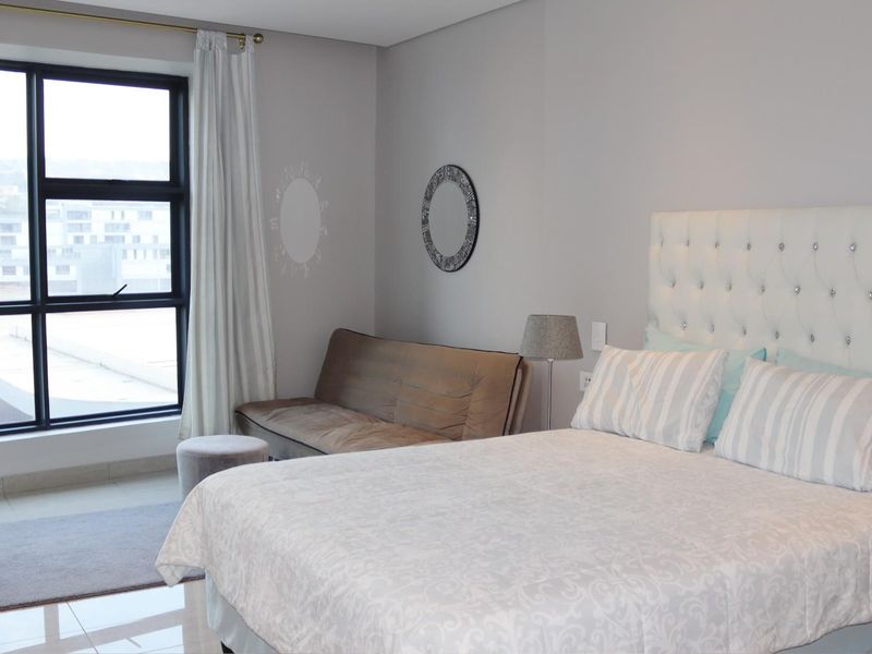 A well-lit bedroom featuring large aluminium-framed windows, neutral wall finishes, and tiled flooring, offering a clean and comfortable living space.