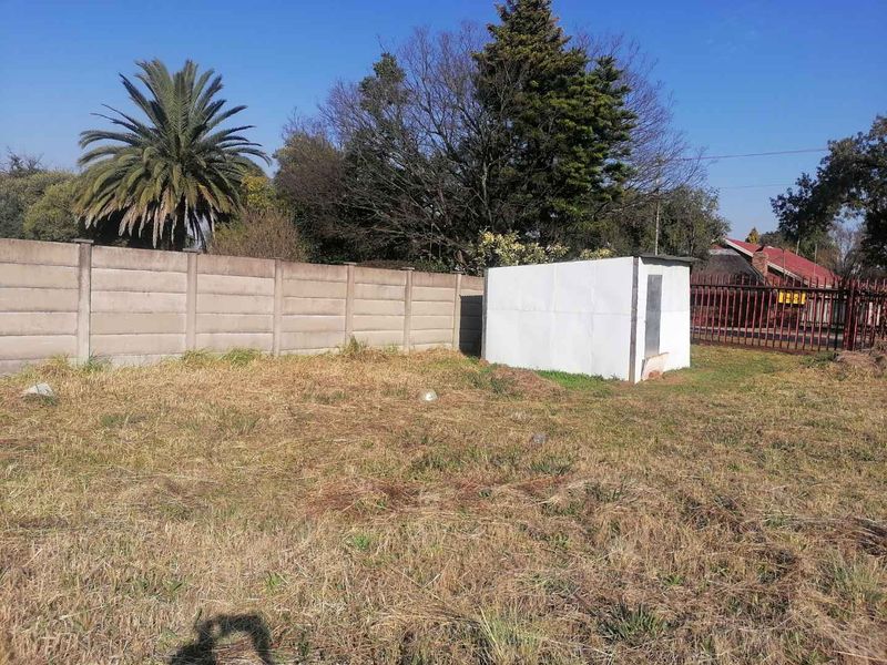 A vacant residential plot featuring a precast concrete boundary wall, metal perimeter fencing, and a small storage shed on the property.
