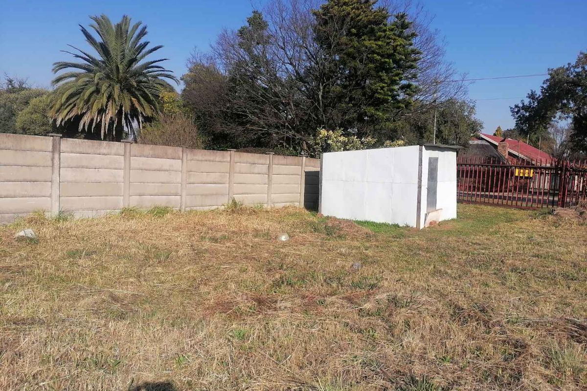 A vacant residential plot featuring a precast concrete boundary wall, metal perimeter fencing, and a small storage shed on the property.