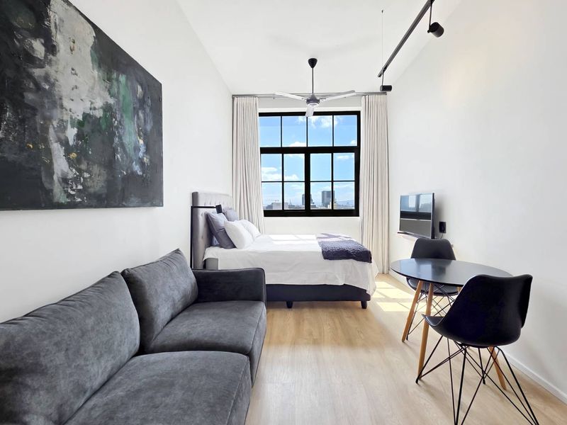 A modern open-plan studio apartment featuring light laminate flooring, industrial-style black-framed windows, and contemporary track lighting with a bright, airy atmosphere.
