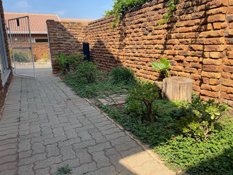 A side garden area featuring a paved walkway, a rustic brick boundary wall, and a metal security gate leading to the exterior.