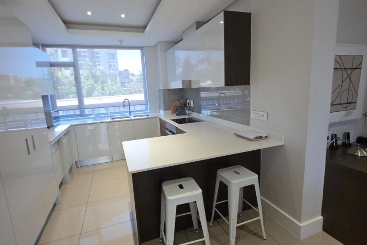 Modern kitchen featuring high-gloss cabinetry, stone countertops, a breakfast bar, and integrated appliances, illuminated by recessed ceiling lighting.