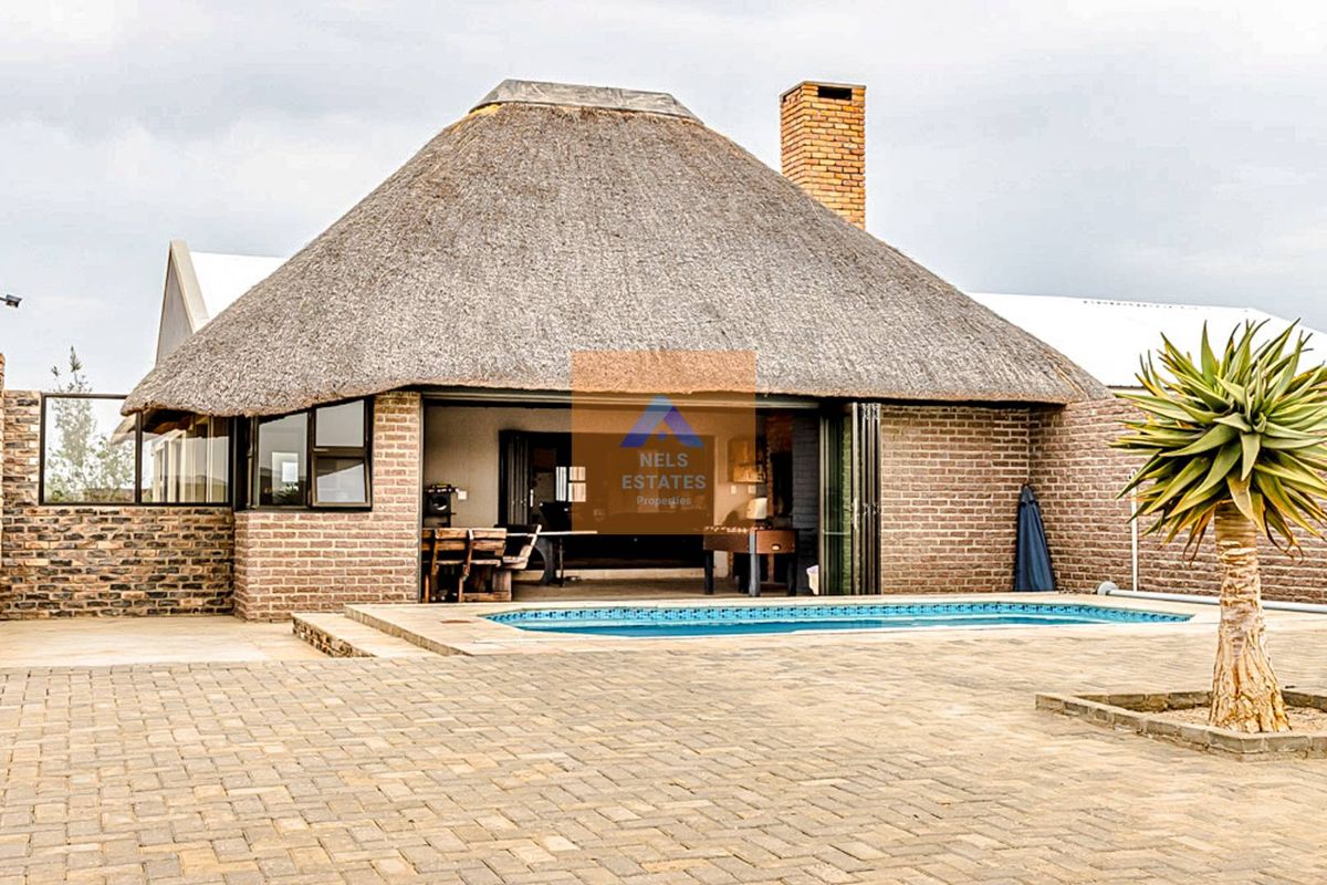 Rear exterior view featuring a thatched roof structure, face brick facade, swimming pool, and paved patio area with large stacking doors.