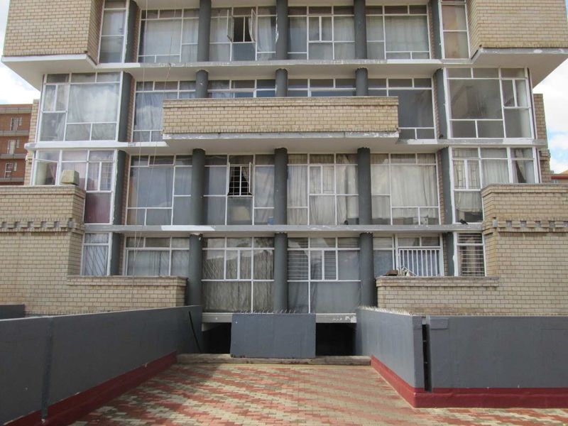 Rear elevation of a multi-story apartment building featuring brick facade, large windows, concrete columns, and a paved ground-level terrace area.