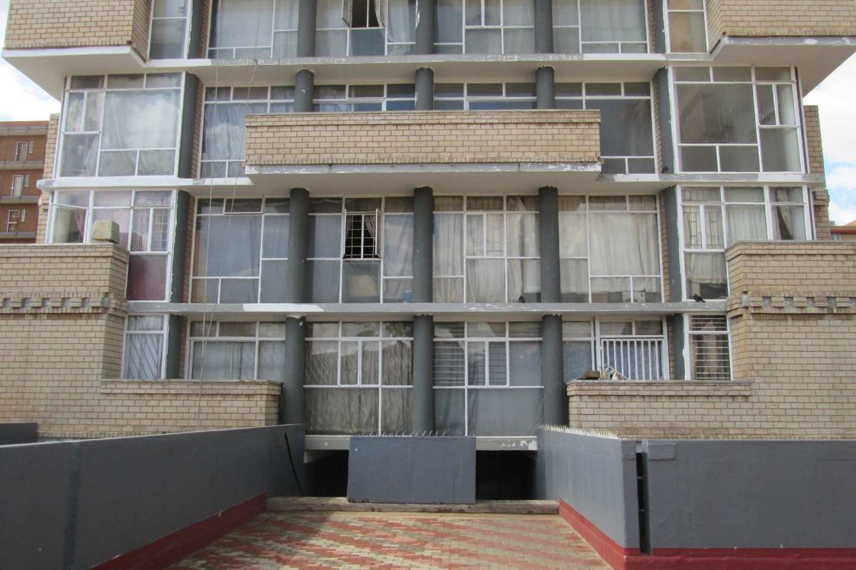 Rear elevation of a multi-story apartment building featuring brick facade, large windows, concrete columns, and a paved ground-level terrace area.