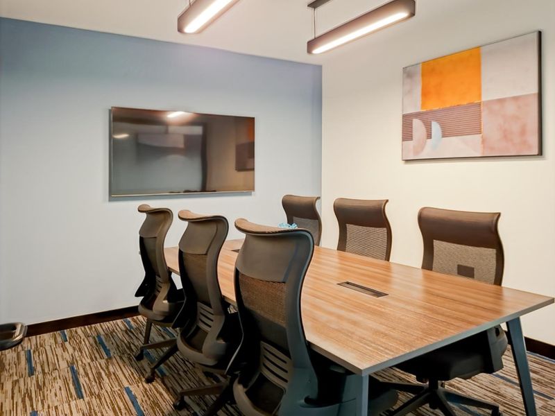 A modern, well-lit office meeting room featuring a professional conference table, ergonomic seating, and a wall-mounted display screen for presentations.