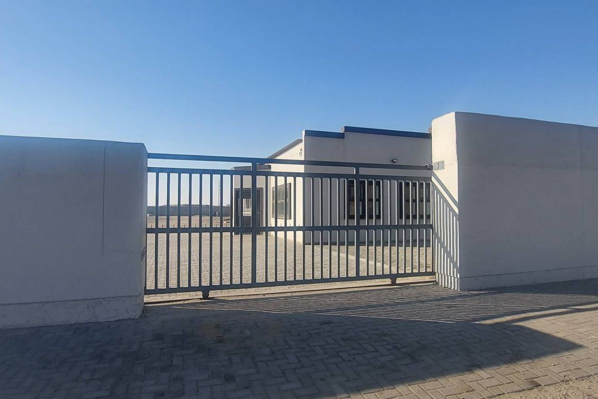 Front view of an industrial property featuring a secure metal sliding gate, perimeter wall, and paved driveway leading to the main building.