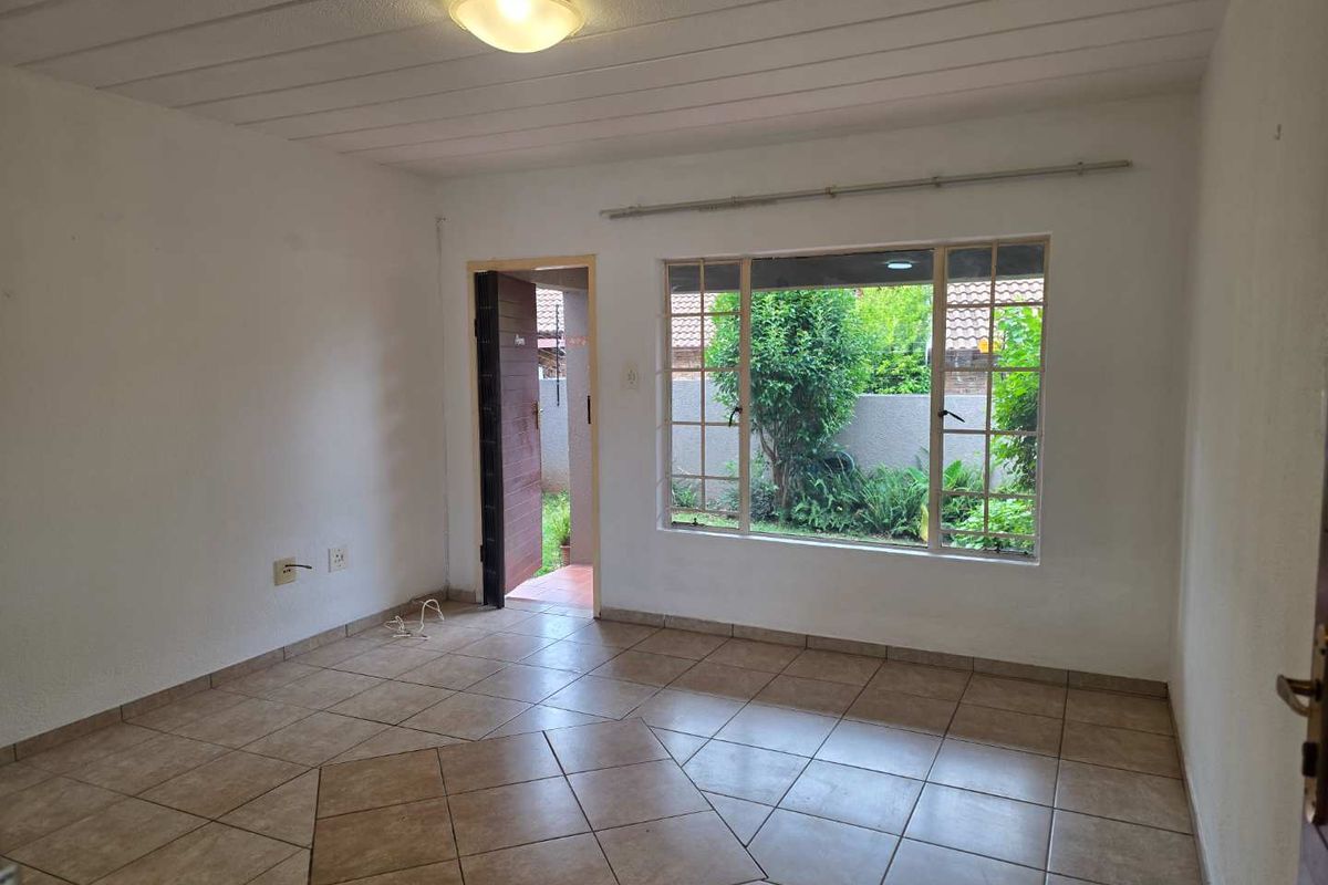 A bright, empty living room featuring tiled flooring, a large window with garden views, and direct access to the exterior.