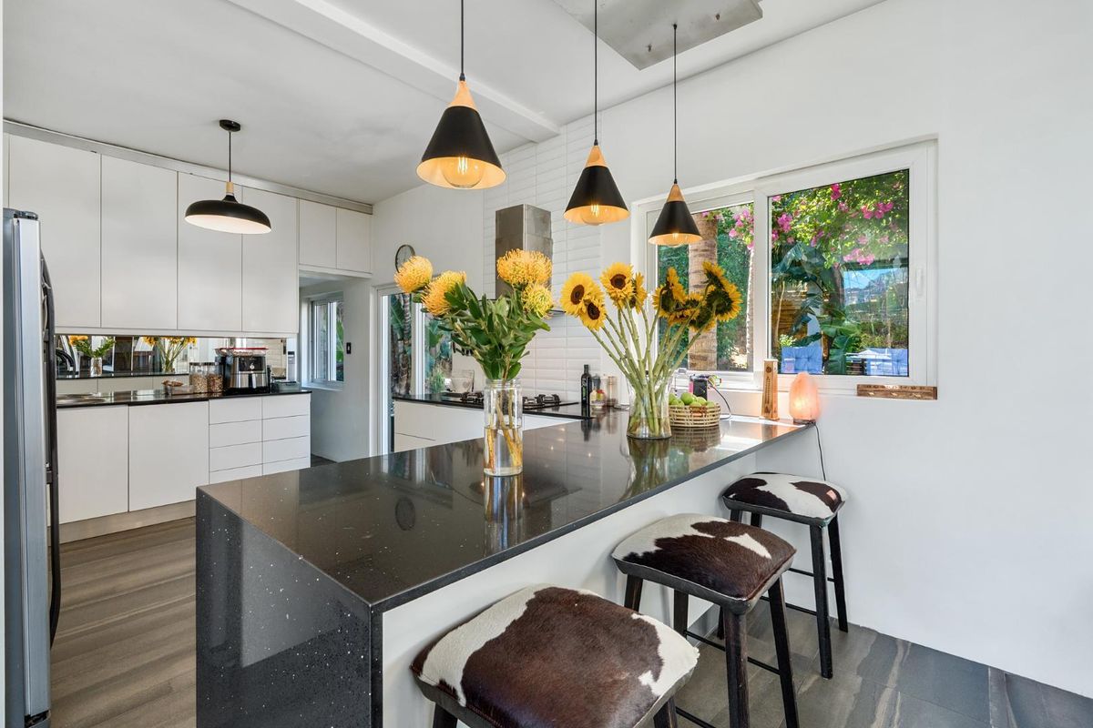 Modern kitchen featuring a black stone breakfast bar, white cabinetry, stainless steel extractor fan, and stylish pendant lighting with a view to the garden.