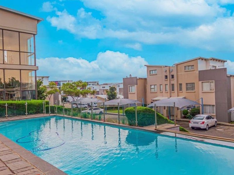 A well-maintained swimming pool area featuring stone paving, a modern glass balustrade, and views of the apartment complex and parking facilities.