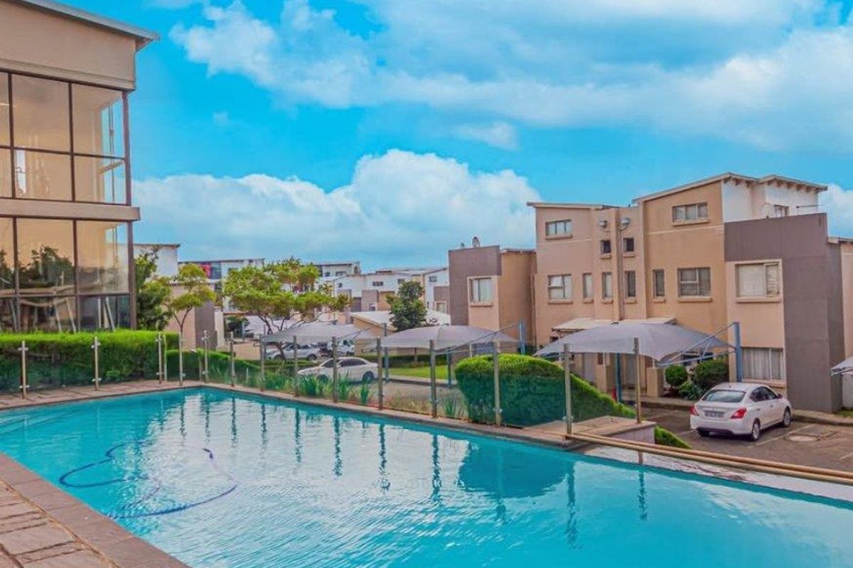 A well-maintained swimming pool area featuring stone paving, a modern glass balustrade, and views of the apartment complex and parking facilities.