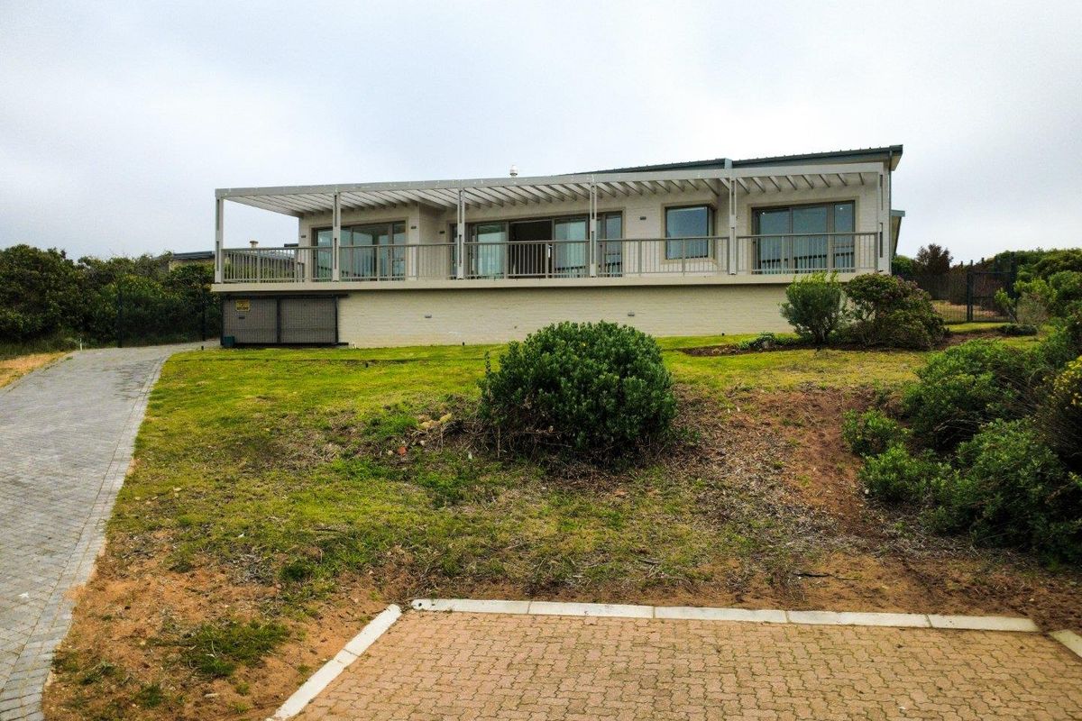 Rear view of a single-story house featuring a large elevated balcony with a pergola, sliding glass doors, and a paved driveway leading to the property.