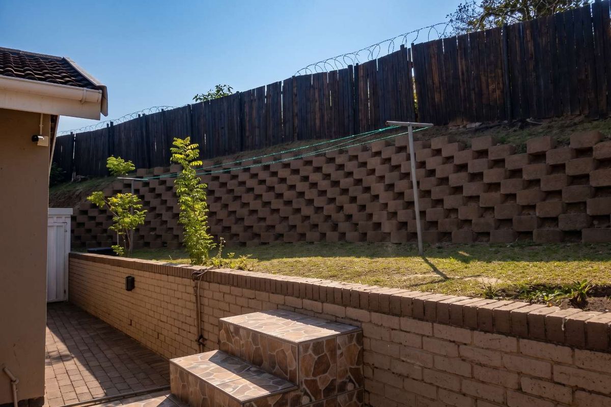 Rear exterior area featuring a multi-level garden with a brick retaining wall, paved patio, tiled steps, and a timber boundary fence with security wire.