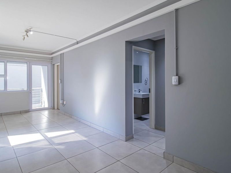 A bright, empty living area featuring neutral tiled floors, grey walls, and access to an outdoor balcony, with a view into the adjacent bathroom.