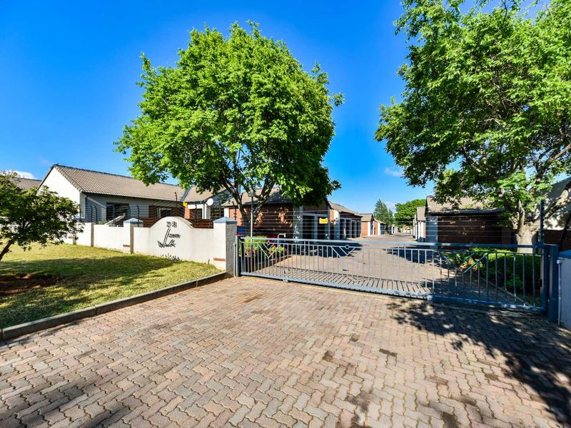 Front entrance view of the townhouse complex featuring a paved driveway, secure gated access, perimeter walling with electric fencing, and established garden areas.