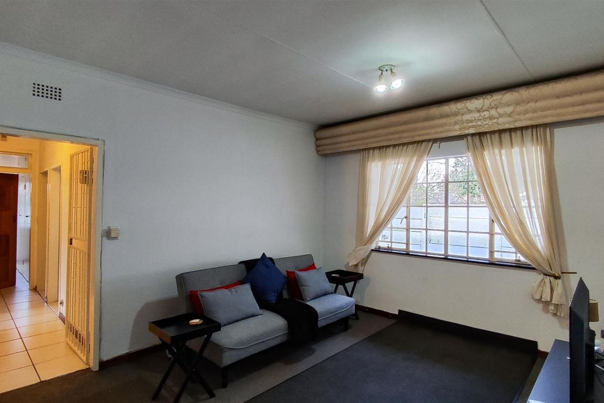 A living room featuring a grid-pattern window with curtains, neutral walls, and access to an adjacent hallway through a security gate.