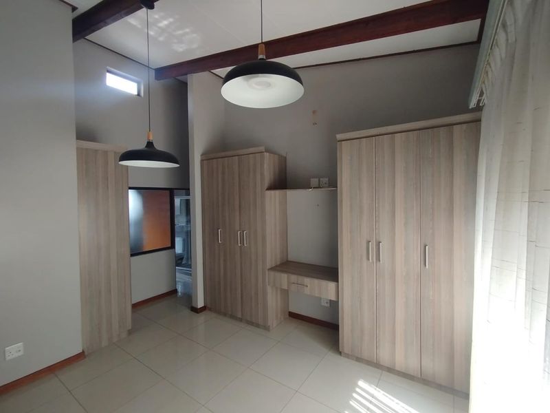 A bright bedroom featuring extensive built-in cabinetry, a dedicated desk area, exposed timber ceiling beams, and neutral tiled flooring.