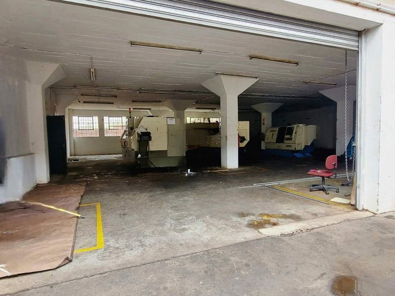 Spacious industrial workshop or garage area featuring concrete flooring, structural support columns, and large industrial windows, suitable for manufacturing or storage operations.