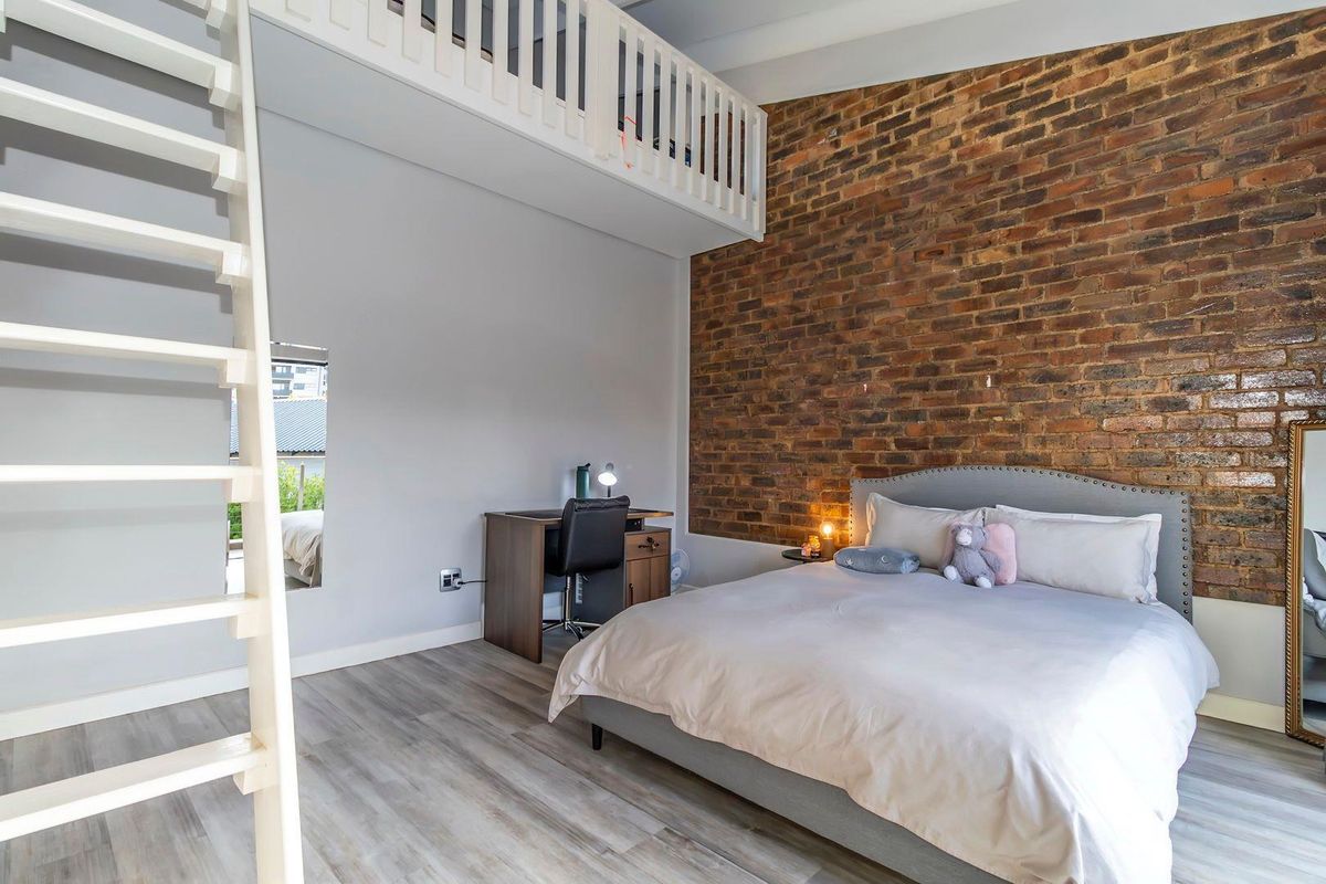 A modern bedroom featuring a striking exposed brick feature wall, light wood-look flooring, and a mezzanine level with a white balustrade.