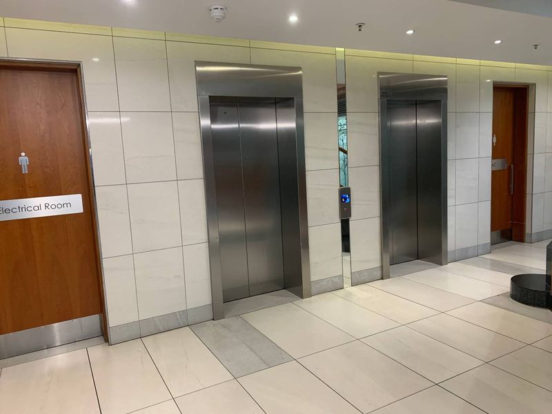 A modern office building hallway featuring stainless steel elevator banks, tiled wall finishes, and clean, well-maintained flooring.