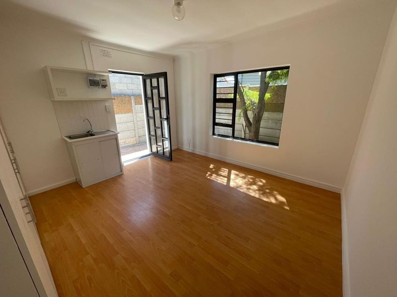 An empty open-plan living area featuring laminate flooring, a compact kitchenette with sink and cabinetry, and direct access to an outdoor space.