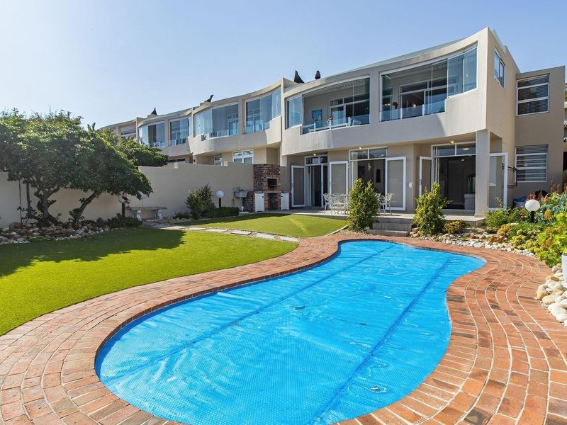 Rear exterior view featuring a swimming pool with brick surround, artificial lawn, built-in braai, and multi-level glass-enclosed balconies.