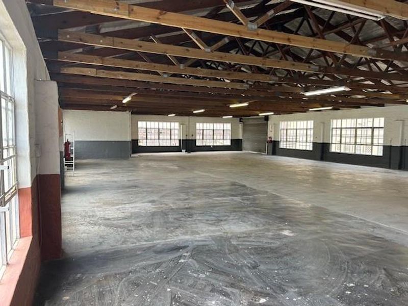 Spacious industrial warehouse area featuring exposed timber roof trusses, concrete flooring, and large steel-framed windows providing ample natural light.