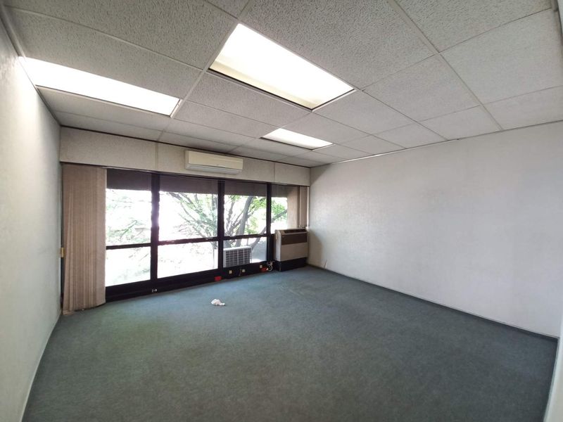 An empty office space featuring a drop ceiling with recessed lighting, wall-mounted air conditioning unit, and large windows providing natural light.
